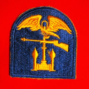May include: A blue embroidered patch with a gold eagle, rifle, and architectural elements. The patch is rectangular with rounded top corners and is set against a red background.