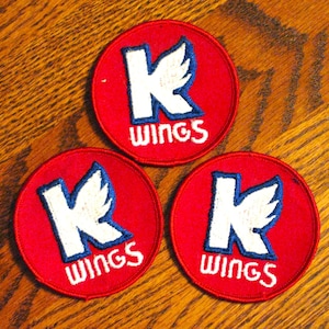 May include: Three red circular patches with a white "K" and wing design, outlined in blue, and the word "WINGS" in white. The patches are on a wooden surface.