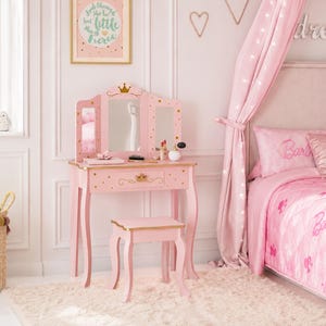 May include: A pink vanity table with a tri-fold mirror, a single drawer featuring a gold crown emblem, and a matching stool. The vanity is decorated with gold star accents and a gold crown at the top center of the mirror frame, standing on curved legs.