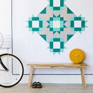 May include: A white wall with a teal and gray quilt hanging on it. A wooden bench sits in front of the quilt with a yellow round pillow on top. A black bicycle with a basket is leaning against the wall.