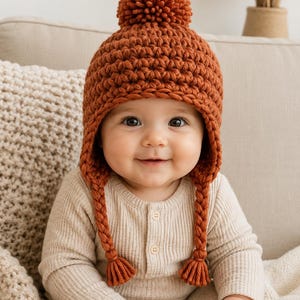 Puede incluir: Un gorro de punto grueso color óxido con un gran pompón en la parte superior, orejeras y dos largas y gruesas trenzas que cuelgan de los lados. El gorro está confeccionado con lana gruesa con un patrón de punto texturizado visible.