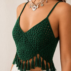 Puede incluir: Un top corto de ganchillo en color verde oscuro con escote en pico, tirantes finos y un dobladillo en punta rematado con borlas de flecos verdes a juego. La prenda está confeccionada con un patrón cuadrado repetitivo y se lleva con una falda blanca.