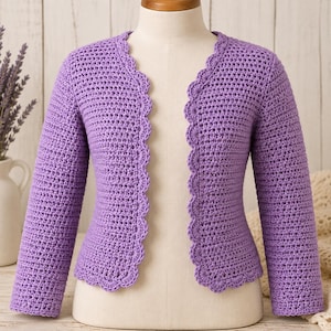 May include: A light purple long-sleeved cardigan made of crocheted yarn. The garment features a scalloped edge detail along the front opening and the bottom hem. It is displayed on a mannequin, showcasing the textured stitch pattern throughout.