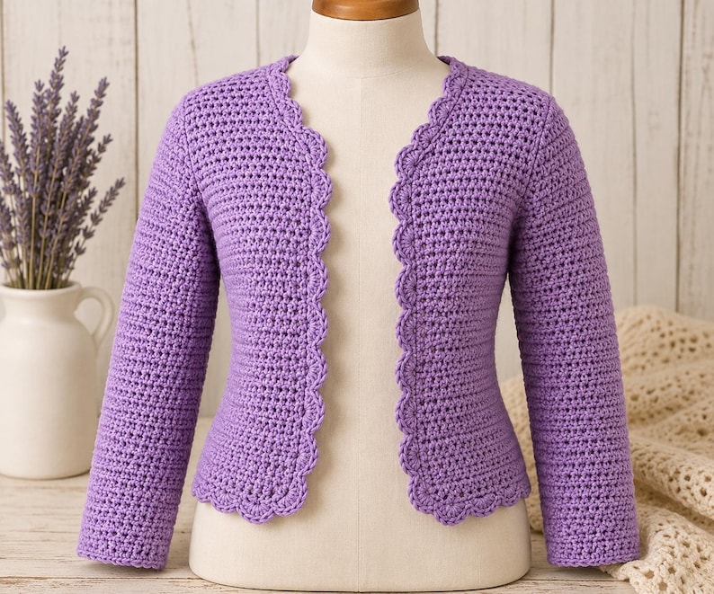 May include: A light purple long-sleeved cardigan made of crocheted yarn. The garment features a scalloped edge detail along the front opening and the bottom hem. It is displayed on a mannequin, showcasing the textured stitch pattern throughout.