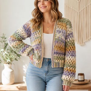 May include: A long-sleeved crochet cardigan featuring a multi-coloured granny square pattern in shades of sage green, lavender, mustard yellow, and cream. The garment has a ribbed hem and cuffs, an open front design, and a straight, cropped silhouette.