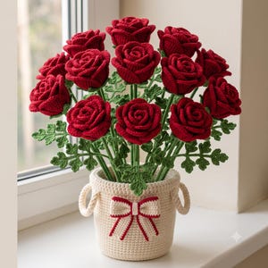 May include: A bouquet of deep red crocheted roses with green crocheted leaves and stems, arranged in a cream-colored crocheted basket. The basket features two side handles and a decorative cream and red bow attached to the front center.