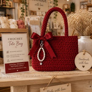 May include: A deep red crochet tote bag featuring a braided handle and a dark red satin ribbon bow. A charm consisting of a string of white faux pearls and two small silver star accents hangs from the center of the bow on the front of the bag.