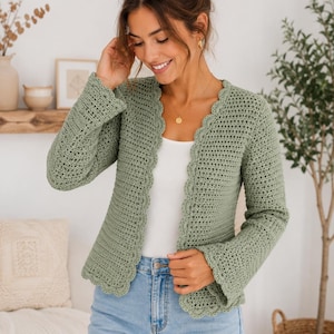 May include: A sage green long-sleeved crochet cardigan featuring a scalloped edge along the front opening and cuffs. The garment is worn over a plain white top and light blue denim jeans, showcasing a textured stitch pattern throughout the piece.
