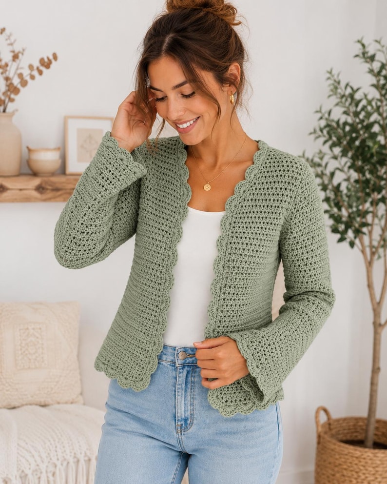 May include: A sage green long-sleeved crochet cardigan featuring a scalloped edge along the front opening and cuffs. The garment is worn over a plain white top and light blue denim jeans, showcasing a textured stitch pattern throughout the piece.