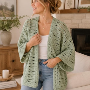 Pode incluir: Um cardigã de crochê verde-sálvia aberto na frente, com um padrão de grade texturizado e repetitivo e mangas largas de três quartos. A peça é usada sobre uma regata branca simples e calças jeans azul-claras, apresentando um caimento solto.