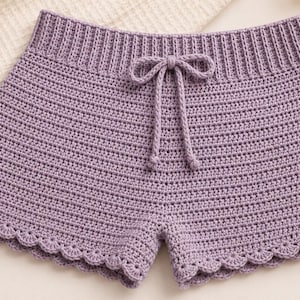 May include: Lavender crochet shorts featuring a ribbed elasticized waistband with a matching drawstring tie. The garment is constructed with a dense horizontal stitch pattern and finished with a decorative scalloped edge along the leg openings.