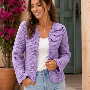 May include: A lavender crochet cardigan featuring long sleeves and a scalloped edge along the front opening and cuffs. The garment is worn over a plain white top and light blue denim jeans, showcasing a textured stitch pattern throughout the piece.