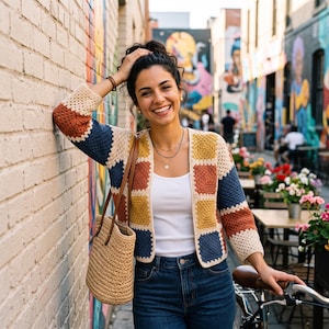 May include: A crochet cardigan featuring a patchwork pattern of square motifs in cream, mustard yellow, navy blue, and burnt orange. The garment has three-quarter length sleeves and an open front, worn over a white tank top and blue denim jeans.