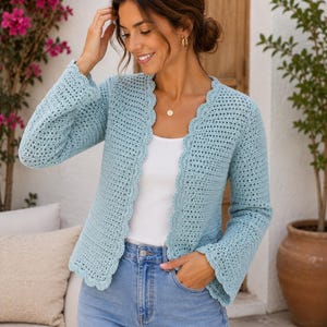 May include: A light blue crochet cardigan featuring long sleeves, a scalloped edge along the front opening, and a textured open-knit pattern. The garment is worn over a plain white top and light blue denim jeans, showcasing a consistent stitch design.