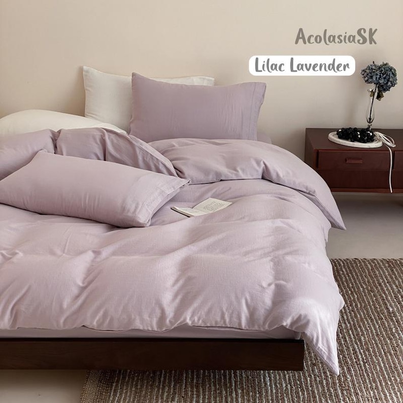 May include: A bedding set in a solid lilac lavender color, featuring a duvet cover and two matching rectangular pillowcases. The text AcolasiaSK and Lilac Lavender is visible in the upper right corner. The bedding is arranged on a bed frame.