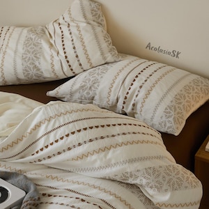 Può includere: Un set di biancheria da letto con due cuscini e un copripiumino in tessuto bianco sporco con motivi geometrici e strisce ricamate in marrone. Il testo AcolasiaSK è stampato in alto a destra. La biancheria poggia su una superficie marrone.