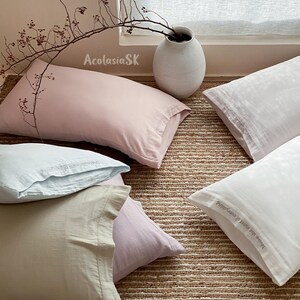 May include: A collection of rectangular pillowcases in soft pastel shades of pink, light blue, beige, and white. Each pillowcase features a stitched border and the embroidered text "Make the most of everything" along the edge of the fabric.