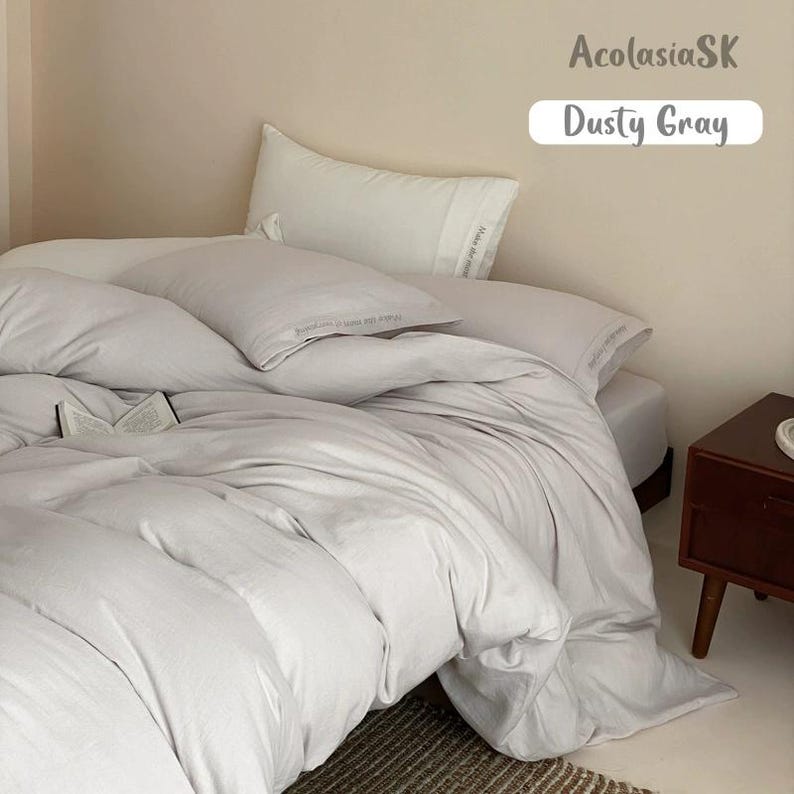 May include: A dusty gray bedding set featuring a duvet cover and two pillows. The bedding has a smooth, solid-colored fabric texture. An open book rests on the duvet. The text AcolasiaSK and Dusty Gray is visible in the upper right corner.