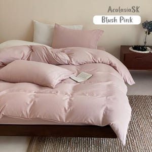 May include: A blush pink bedding set featuring a duvet cover and two matching pillowcases. The bed is made with a white pillow tucked behind the pink one. An open book rests on the duvet. Text in the image reads AcolasiaSK and Blush Pink.