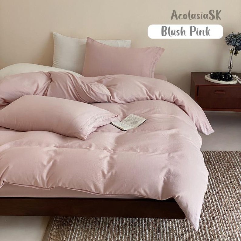 May include: A blush pink bedding set featuring a duvet cover and two matching pillowcases. The bed is made with a white pillow tucked behind the pink one. An open book rests on the duvet. Text in the image reads AcolasiaSK and Blush Pink.