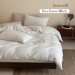 May include: A bed set featuring a cream white duvet cover and matching pillowcases. The bedding has a soft, textured appearance. An open book rests on the duvet. The text AcolasiaSK and Pure Cream White is visible in the upper right corner.
