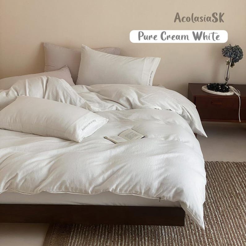 May include: A bed set featuring a cream white duvet cover and matching pillowcases. The bedding has a soft, textured appearance. An open book rests on the duvet. The text AcolasiaSK and Pure Cream White is visible in the upper right corner.