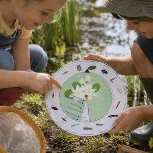 Op de afbeelding: Twee kinderen bekijken een ronde educatieve kaart met insectenillustraties en een waterlelie-diagram. Een klein schoolbord met de tekst "Look closely, Ask questions, Respect nature" is zichtbaar. Een net en een bak met water staan op de voorgrond.