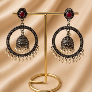 May include: A pair of oxidised silver-toned earrings featuring a circular frame with intricate detailing, a central maroon stone stud, and a dangling bell-shaped jhumka element. The design is accented with small white beads hanging from the bottom.