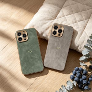 Puede incluir: Dos fundas de teléfono personalizadas. Una es verde apagado con las iniciales "TQ" y la otra es gris con "GD". Ambas fundas tienen anillos de cámara y bordes dorados. Las fundas están sobre una superficie de madera con una almohada y arándanos en el fondo.