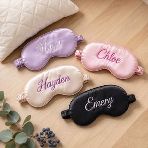 May include: Four satin sleep masks with elastic headbands, each featuring custom embroidery. The lavender mask reads Maya, the pink mask reads Chloe, the champagne mask reads Hayden, and the black mask reads Emery in elegant script lettering.