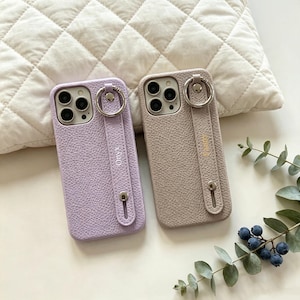 May include: Two textured phone cases with integrated finger ring straps. The left case is light purple with the text Onyx, and the right case is beige with the text Jenny. Both feature silver-toned hardware and cutouts for the camera lenses.
