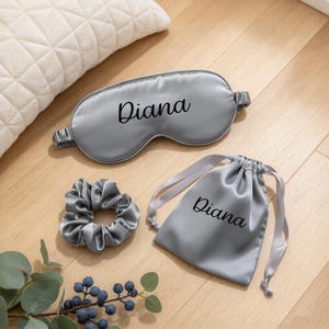 May include: A silver-gray satin sleep mask, a matching scrunchie, and a drawstring pouch. The sleep mask and the pouch both feature the name Diana printed in a black cursive script. The set is made of a smooth, shiny fabric with a consistent color.