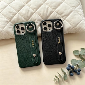 May include: Two textured phone cases, one dark green and one black, each featuring a vertical strap with a silver metal ring and a silver stud. The green case is personalised with the name Chloe in gold, and the black case with the name Rosie.