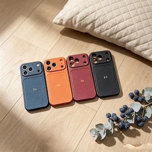 Pode incluir: Quatro capas de telefone de couro em azul, laranja, bordô e preto, cada uma com iniciais diferentes. As capas estão dispostas sobre uma superfície de madeira, com uma almofada ao fundo. Um ramo de bagas e folhas está em primeiro plano.