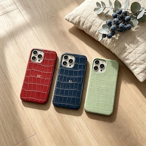 Puede incluir: Tres fundas para teléfono en rojo, azul marino y verde claro, cada una con textura de piel de cocodrilo. La funda roja tiene "HP" en relieve, la azul marino "KL" y la verde claro "FQ". Sobre una superficie de madera.