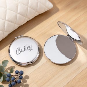 Puede incluir: Dos espejos de bolsillo redondos de metal plateado. Uno está cerrado y tiene el nombre Bailey grabado en letra cursiva en la tapa. El segundo espejo está abierto, mostrando una superficie reflectante y un mecanismo de bisagra lateral.