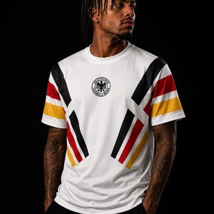 Op de afbeelding: Een wit T-shirt met korte mouwen en een ronde hals, voorzien van zwarte, rode en gele diagonale strepen op de mouwen en de romp. In het midden van de borst staat een rond zwart-wit embleem met de tekst Deutscher Fussball-Bund.