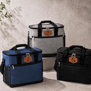 May include: Three insulated cooler bags in grey, blue, and black, each featuring black handles, a front zippered pocket, and a brown leather-like patch with a personalized name or initial. The bags have a rectangular shape and silver zippers.