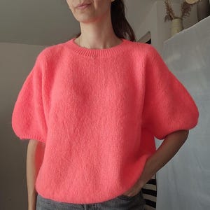Peut inclure: Un pull en tricot rose néon vif doté d'une encolure ronde et de manches courtes bouffantes volumineuses. Le vêtement est confectionné avec un fil texturé duveteux et présente une silhouette décontractée et ample qui tombe à la taille.