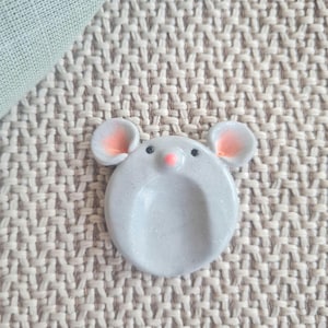 Könnte beinhalten: Eine kleine, kreisförmige, hellgraue mausförmige Ablageschale mit einem dezenten Glitzer-Finish. Die Schale hat zwei abgerundete Ohren mit zartrosa Mitten, zwei kleine schwarze Augen und eine winzige rosa Nase über einer glatten, konkaven Vertiefung.