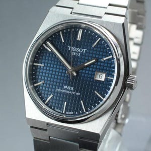 May include: A stainless steel Tissot PRX Powermatic 80 watch with a blue dial. The watch features a date window at the 3 o'clock position and a silver-tone bracelet. The dial has a textured pattern and the watch is marked "Swiss Made".