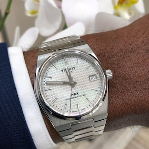 Puede incluir: Un reloj Tissot PRX Powermatic 80 plateado con esfera blanca estampada y visualización de fecha a las 3. El reloj tiene una pulsera de acero inoxidable. Fondo borroso con flores blancas.