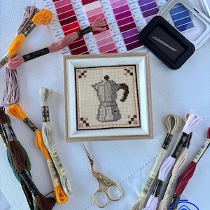 May include: A framed cross-stitch embroidery of a moka pot coffee maker, surrounded by various colorful embroidery floss skeins, a needle minder, a color chart, and gold-toned stork-shaped embroidery scissors on a white fabric background.