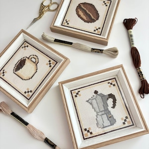 May include: Three square wooden frames containing cross-stitch art of a coffee bean, a coffee mug, and a moka pot. Gold embroidery scissors and three skeins of embroidery floss in beige, light tan, and dark brown are arranged around the frames.