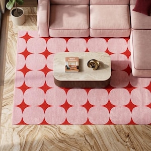 May include: A rectangular area rug featuring a repeating pattern of light pink rounded squares separated by red four-pointed star shapes. The rug is positioned beneath a light-colored coffee table with a book titled Retro Interiors and a gold sculpture.