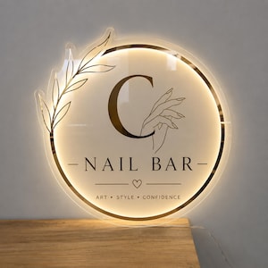 Op de afbeelding: Een rond LED-acrylbord met een goudkleurige letter C, een lijntekening van een hand en de tekst NAIL BAR met daaronder de woorden ART, STYLE, CONFIDENCE. Een gouden takontwerp accentueert de linkerkant van de ronde omlijsting.
