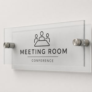 May include: A rectangular clear acrylic sign with a frosted backing, mounted with two silver metal standoffs. The sign features a black icon of three people at a table, with the text MEETING ROOM and CONFERENCE printed in black sans-serif font.
