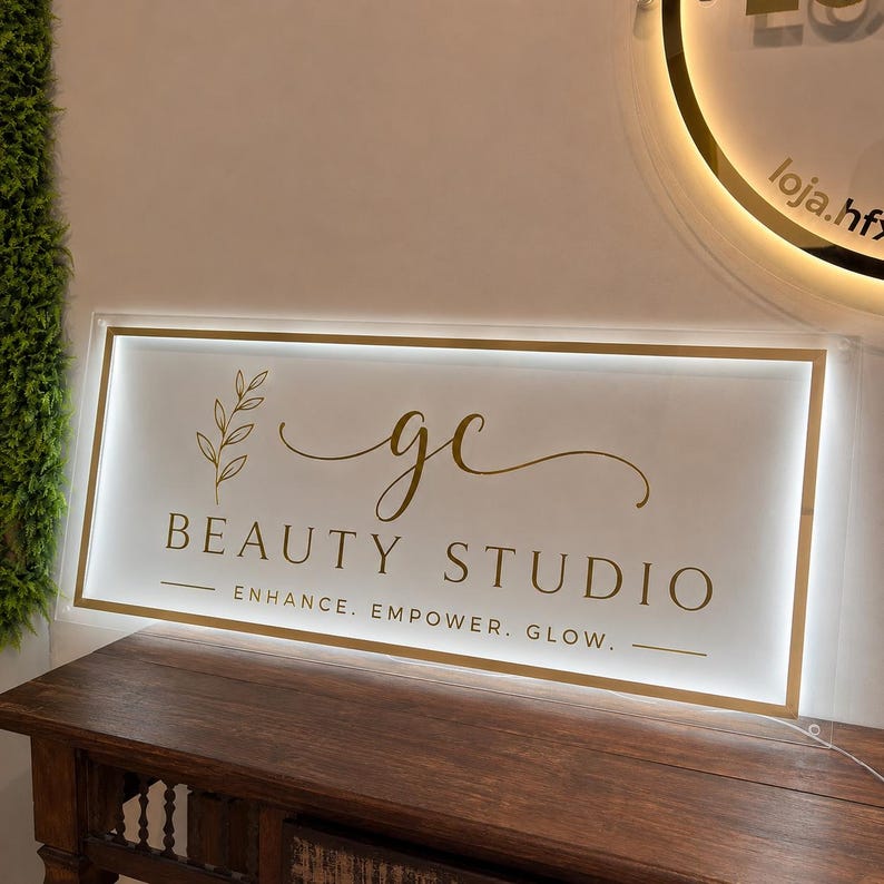 May include: A rectangular, illuminated acrylic sign featuring gold lettering and a leaf graphic. The text reads gc BEAUTY STUDIO and ENHANCE. EMPOWER. GLOW. The sign is backlit with a white glow, framed by a gold border, and mounted on a wooden surface.