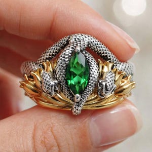 May include: A ring featuring a central marquise-cut green gemstone held by two silver-toned textured snake figures. The band incorporates gold-toned metallic elements shaped like stylized leaves or petals, creating a layered design around the stone.