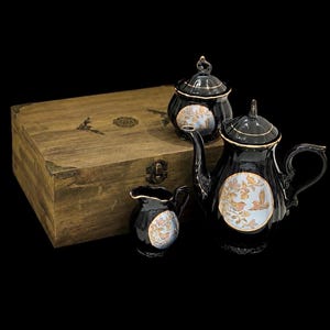 Puede incluir: Un juego de té de cerámica negra que incluye una tetera, un azucarero con tapa y una jarra de leche, decorados con medallones circulares que muestran pájaros sobre ramas. El conjunto se presenta junto a una caja de madera rectangular.